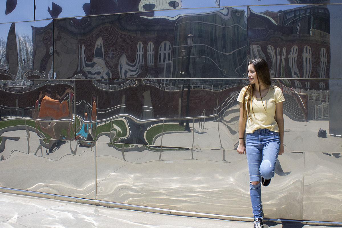 Angela poses in front of a mirror building with skinny jeans, sneakers and a yellow shirt.