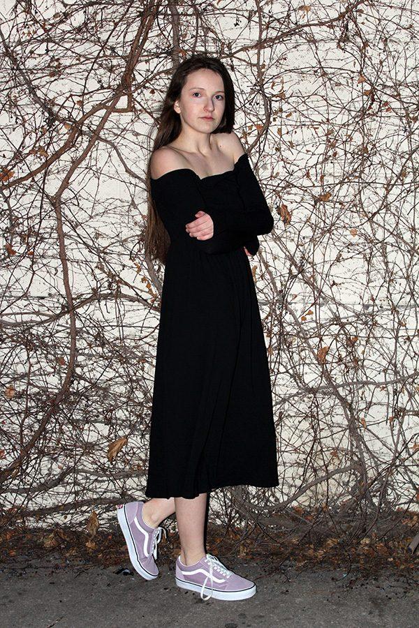 Wallace stands against a wall covered in vines wearing a black, strapless dress from Urban Outfitters with lavender Vans.